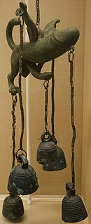 A winged metal penis with two back legs and a tail is suspended on a chain. Five bells are suspended from the head of the penis, one from each wing and one from each back foot