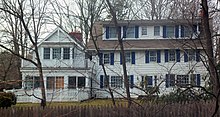 A two-story, five-bay white wooden house with blue shutters and pent-roofed dormers piercing its gabled roof seen over a fence and through some tree branches. It has a more modern wing on the left