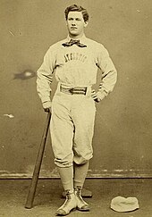 A baseball player is standing, facing the camera, with the top of a baseball bat in his right hand, and the bottom resting on the ground