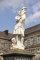 Juana de Arco o Jeanne d'Arc au sacre, fundición, fecha no precisa, estatua pedestre con armadura, portando un estandarte; en el adro de la iglesia de Santa María de Tinchebray, Orne, Baja Normandía, Francia.[17]​