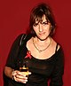 Tracey Emin, holding a wine glass filled with a translucent, peach-colored liquid. She is wearing a black top, with a red ribbon attached.