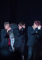 Three men dressed in black suits perform on stage with one hand covering their face partially.