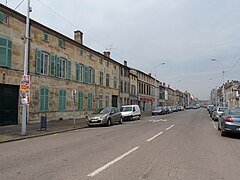 Large rue avec des voitures stationnées de chaque côté, encadrée d'immeubles.