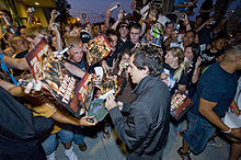 A crowd of people is all looking towards a man at the center who is signing a hat. The crowd is attempting to hand him posters to sign and others are taking pictures using cameras and cell phones.