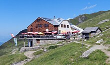 Ansicht auf die Karl-von-Edel-Hütte, an einem mit Gras bewachsenden Berghang, vorn ein aus Stein erbautes Gebäude, die Terrasse mit Pfeilern über den Hang gebaut; weiter hinten ein verputztes Gebäude; im Hintergrund zwei Masten mit Fahnen