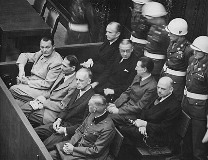 Eight men seated in two rows with a line of guards behind them