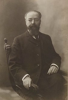 Photo sépia d'un homme assis, les jambes croisées, en costume trois pièces avec cravate ; portant une barbe, il a les cheveux grisonnants et dégarnis