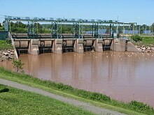 A concrete and steel structure crosses the river. The water is brown on the near side of the structure, and blue on the far side