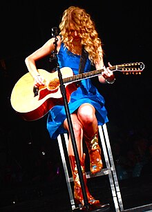 Taylor Swift, donning a royal blue sundress and playing a wooden acoustic guitar