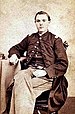 Young white man sitting on a chair with his legs crossed and his right arm resting on a table beside him. He is wearing light colored pants and a military jacket buttoned at the top only with a vest underneath.