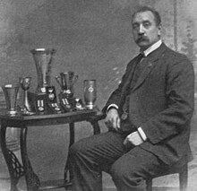 Black and white photo of a middle-aged man wearing a suit with a white collar shirt, sitting a table including multiple trophies and medallions