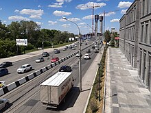 Photographie de la rue Bolchévique à Novossibirsk. Il y a trois voies dans un sens, quatre dans l'autre. Du côté droit, on observe un bâtiment gris dont on ne peut déterminer la fonction, tandis qu'à gauche se trouve un bois. Des lampadaires longent la route, reliés entre eux par une multitude de fils électriques. Le trafic n'est pas excessif, et en arrière-plan, on observe la construction de deux grandes tours, avec d'imposantes grues.