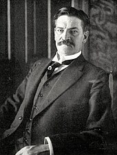 Photograph of a moustached middle-aged man in a dark suit and waistcoat, sitting in a chair while looking at the camera