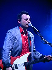 Photograph of James Dean Bradfield singing into a mic while playing an acoustic guitar