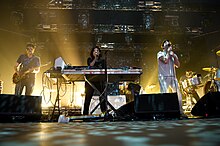 A man playing guitar, a woman playing the keyboard, and a man singing on stage