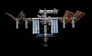 A forward view of the International Space Station with limb of the Earth in the background. In view are the station's sixteen paired maroon-coloured main solar array wings, eight on either side of the station, mounted to a central integrated truss structure. Spaced along the truss are ten white radiators. Mounted to the base of the two rightmost main solar arrays pairs, there are two smaller paired light brown-coloured ISS Roll-out Solar Arrays. Attached to the centre of the truss is a cluster of pressurised modules arranged in an elongated T shape. A set of solar arrays are mounted to the module at the aft end of the cluster.
