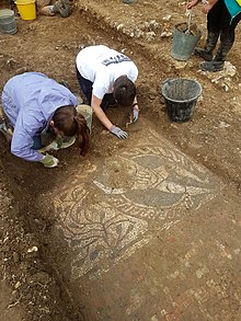 Two people kneeling in front of a mosaic, trowels in hand.