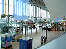 Inside a terminal, with a multi-story glass wall to the left; the open space is filled with signs, chairs, small store and amenities