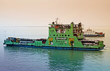 A green ferry boat named Shah Ali with blue and white text on the side sails across a large body of water. Buses and trucks are lined up on the lower deck of the ferry with people. There are also people on the three upper decks. Advertisement text on the side of the ferry reads, "ইস্পাহানি" (Ispahani), and "মির্জাপুর" (Mirzapur)