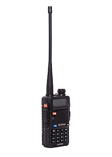 This image shows a Baofeng UV-5R, a small black handheld transceiver. It has a few buttons on the side and numerical buttons and a small LCD display on the front.