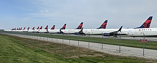 Many in line airplanes with the Delta Air Lines logo on the tail, parked on pavement behind a fence.