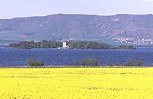 A field of yellow flowers in the foreground, with a dark blue lake beyond. A wooded island in the lake has a white structure of two storeys at centre and there are green and brown hills beyond. There is a small cluster of houses on the distant hill slope at right.