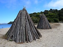 Two Miwok bark houses, cone shaped, on the beach with water and trees in the background