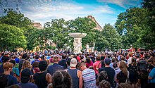 Hundreds of people stand together listening to a man speaking from an outdoor stage. In the center of the crowd is a large statue with a paper sign affixed to it with tape; the sign reads: "D.C. Loves You Orlando."