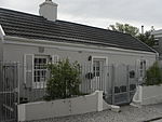 Free-standing single storey vernacular cottage on street edge; double pitched roof with aluminium strip sheeting and end bargeboards; possibly original sash windows (12 panes) with partly original glass; stable door with small-paned glazing; low stoep Type of site: House Current use: House.