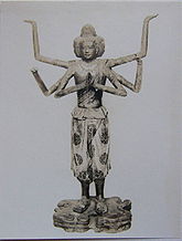 Front view of a standing statue with six arms and three faces. One pair of hands is joined in front of the body with the palms facing each other as if praying. A second set of arms is bend to the sides with the palms of the hands pointing upwards. Also the third pair of arms is bend somewhere between the positions of the other pairs of arms. The three faces point to the fron and both sides of the statue. The statue is embellished with sculpted and painted wide three-quarter length trousers and sandals. Black and white picture.