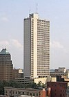 A 34-story whitish office building stands against a largely clear sky. The tower is boxy and mostly windows interspersed with concrete. To the left stands a lower, darker tower with a green roof.