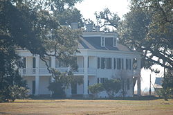 Three story tall large white house with fields in the foreground and trees in the background.