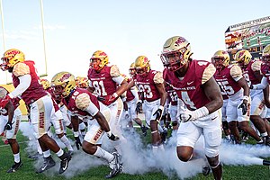 Football players dressed in maroon and gold run on the football turf field with smoke surrounding them.