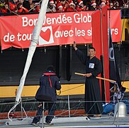 Debout sur le pont, en kimono noir et hakama noir, Kiraishi salue la foule. Un sabre de bois est passé dans sa ceinture.