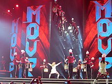 Backshot of Madonna dancing dressed in a white uniform. Several dancers dressed as a marching band are suspended on mid-air.