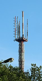 Rising from above a tree, a tall and thick red candelabra tower with four arms bearing different types of broadcasting antennas. One has red and white panel antennas.