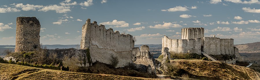 The ruins of a castle in grey limestone dominating the landscape. The River Seine is in the background. The castle's keep protrudes above the walls of the inner bailey on the right, with a bridge leading up to the bailey's entrance. To are ruins of the wall enclosing the outer bailey; a tower stands taller than the ruined walls.