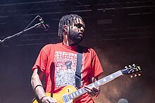 Malani shown in upper body shot, his head turned to his left away from a microphone on a stand. He wears a red tee-shirt with partly obscured, white writing "The person wearing this shirt stands against sexual assault and demands a change". His hair hangs in dreadlocks and he sports a beard. His left hand is on the neck of his guitar and he strums with his right