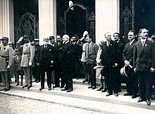 Photographie noir et blanc. Sur un perron une assemblée de personnages, du début du XXe siècle, en tenue de ville pose.