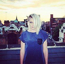 Blonde haired young woman with noticeable eye make up wearing a blue top against an urban rooftop background and orange sky