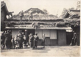 A crowd gathers in front of a detached house in a town to watch thatchers at work. The house frontage lies directly on the street, and consists of a row of shoji (with one solid panel at the end, presumably for a to-bukuro).