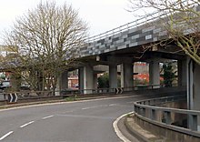 View of a slip road bending round to the right, passing under the concrete-supported viaduct of the main carriageway, decorated with grey tiles