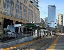 Station platform and canopy with a white tram with orange and red stripes waiting to depart and various passengers visible.