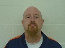 Photo of inmate John Eric Armstrong wearing standard uniform of inmates in the Michigan Department of Corrections consisting of a white undershirt and a dark blue button up shirt with an orange stripe on each shoulder