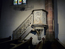 photographie en couleur montrant une petite tribune octogonale en grès gris adossée à un pilier en grès rose et accessible par une escalier doté d’une balustrade en pierre décorée de motifs géométriques