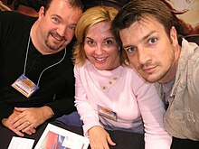 a blonde woman in a pink shirt sits between a man in a black shirt and a goatee (left) and a man with brown hair wearing tan (right)