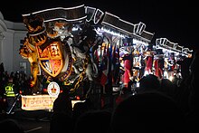 A large vehicle lit by multi-coloured lamps and carrying people in brightly coloured coustumes in front of a large crowd on a darkened street.