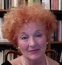 Close-up portrait of Bakró-Nagy, a red-haired woman wearing earrings and a necklace, sitting on a chair in front of a bookcase.