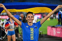 Yaroslava Mahuchikh, maillot bleu avec écrit "Ukraine", brandit un drapeau ukrainien au-dessus d'elle en souriant.