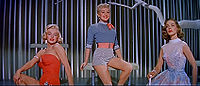 Monroe in How to Marry a Millionaire. She is wearing an orange swimsuit and is seated next to Betty Grable, who is wearing shorts and a shirt, and Lauren Bacall, who is wearing a blue dress.